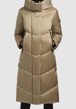 Pitkä beige toppatakki, jossa on korkea kaulus, vinoruutukuvio, sivuvetoketjutaskut ja kädet taskuissa, päällä mustat housut.