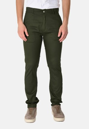 Parte inferiore del corpo di una persona che indossa pantaloni slim fit verde scuro, sneakers beige e una camicia bianca infilata, su sfondo semplice.