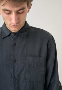 Schwarzes Hemd mit Knöpfen aus weichem Stoff, das eine Brusttasche, einen klassischen Kragen und einen geraden Schnitt mit subtilen Nähdetails aufweist.