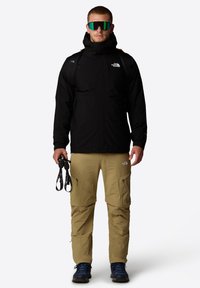 Zwarte waterdichte jas met een capuchon, gecombineerd met lichtbruine cargopants met ritsen. Model houdt zwarte banden vast en draagt een zonnebril met groene tint.