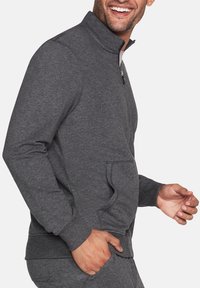 Sudadera gris con cremallera y alto cuello, con bolsillos laterales y puños acanalados, fabricada con un tejido suave y texturizado.