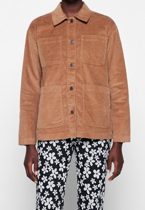 Person iført en lysebrun kordfløjlsjakke med knapper og fire forlommer foran samt sorte bukser med hvidt blomsterprint.