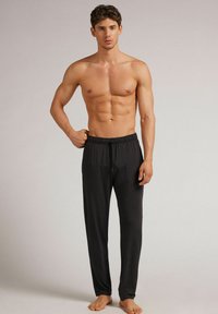 Schwarze Jogginghose aus leichtem Stoff, mit entspannter Passform und schmal zulaufenden Beinen, barfuß von einem Modell ohne Shirt getragen.