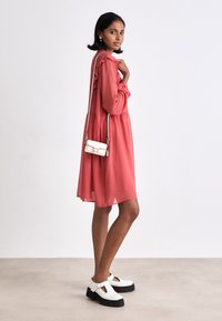 Abito rosa cipria con maniche a volant e un design fluido, abbinato a scarpe con platform bianche. Una piccola borsa a tracolla bianca pende dalla spalla.