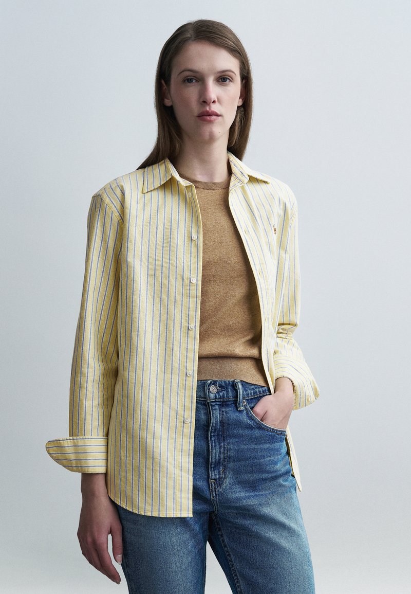 Chemise à rayures jaunes et blanches boutonnée, portée sur un pull marron, associée à un jean bleu ; elle présente un col classique et des manches retroussables.