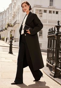 Lång svart kappa med en figursydd krage, i kombination med vida mörka jeans och spetsiga svarta klackar. Tillbehöret består av en liten svart kuvertväska.