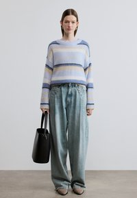 Pull en laine à rayures bleu clair, crème et bleu foncé ; associé à un denim délavé ample. Sac fourre-tout noir et chaussures beiges.