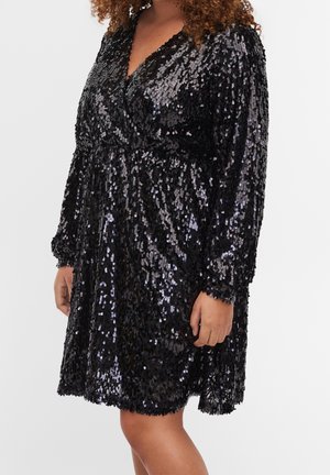 Femme portant une robe noire longueur genoux couverte de sequins brillants, avec des manches longues et un décolleté en V.