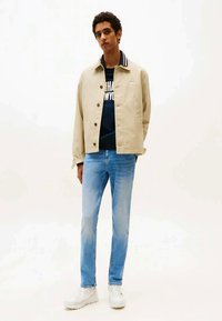 Young man wearing a beige jacket, navy graphic sweatshirt, light blue jeans, and white sneakers, standing against a plain white background.