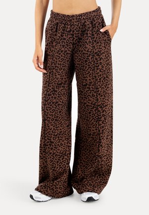 Person iført brune bukser med vide ben med sort leopardprint, elastisk linning, sidelommer og hvide sneakers.