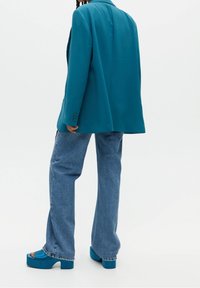 Blazer oversize bleu sarcelle à épaules structurées, associé à un jean évasé bleu clair et des sandales compensées épaisses assorties. Texture de tissu lisse.