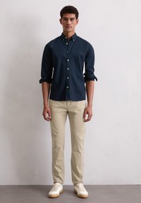 Man in een marineblauwe overhemd met opgerolde mouwen, beige broek en witte sneakers, staande tegen een effen lichte muur.