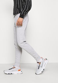 Osoba ubrana w szare dresy i białe sneakersy z pomarańczowymi i czarnymi akcentami idąca na jednolitym białym tle.