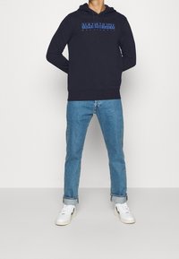 Felpa navy con cappuccio e cordino, caratterizzata da testo stampato blu sul petto. Abbinata a jeans azzurri chiari e sneakers bianche.