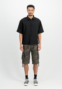 Melns īsās piedurknes krekls ar pogām, apvienots ar zaļiem kamuflāžas šortiem ar sānu kabatām un melniem sneakeriem ar baltām auklām.