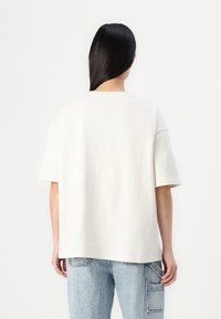 Camiseta blanca de algodón con mangas cortas, un escote redondeado y un ajuste relajado. Llevada con jeans de mezclilla azul claro que tienen un bolsillo de parche.