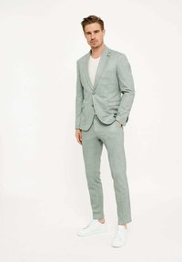 Costum verde deschis dintr-un material texturat, cu rever ascuțite, asortat cu un tricou crem și adidași albi. Croială adaptată și design modern.