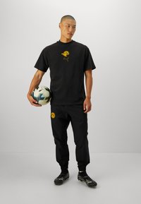 Sort atletisk outfit med en kortærmet t-shirt med gult logo og sorte træningsbukser med gul emblem. Holder en fodbold.
