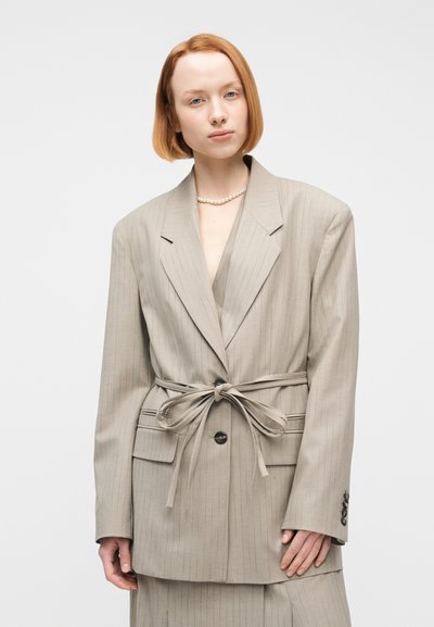 Γυναίκα με κοντά κόκκινα μαλλιά που φοράει μια μπεζ oversized σακάκι με λεπτές ρίγες, δεμένη με ζώνη και ένα κολιέ από πέρλες, σε λευκό φόντο.