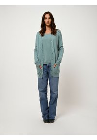 Ljusblå stickad cardigan med knappar, ett par blå jeans och mörka skor. Cardiganens har fickor och en mjuk textur.