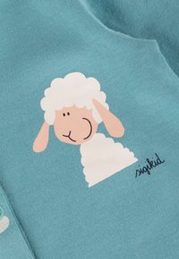 Türkiser Stoff mit einem Schafdesign, das einen weißen, strukturierten wolligen Körper und ein beige-farbenes Gesicht zeigt, ergänzt durch schwarze Gesichtszüge und eine Signatur.