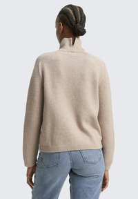 Beżowy, teksturowany sweter z wysokim kołnierzem i prążkowanymi mankietami, widziany z tyłu. W komplecie ze spodnimi dżinsowymi o jasnym odcieniu.