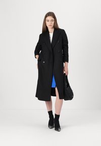Cappotto nero doppiopetto con grandi bottoni, maniche lunghe, tasche laterali, abbinato a un vestito blu, borsa in tessuto nero strutturato e stivaletti neri.