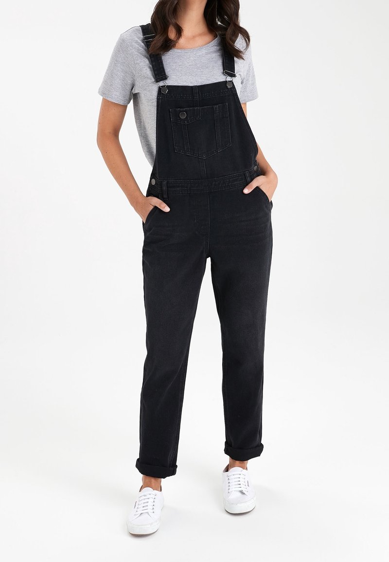 Femme portant une salopette en denim noir avec les revers roulés, un t-shirt gris clair à manches courtes et des baskets blanches, debout les mains dans les poches.