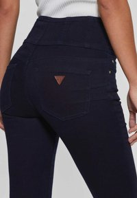 Vysoké tmavě modré džíny s texturovanou látkou, které mají hnědý trojúhelníkový logo na zadní kapse a zlaté kovové akcenty.