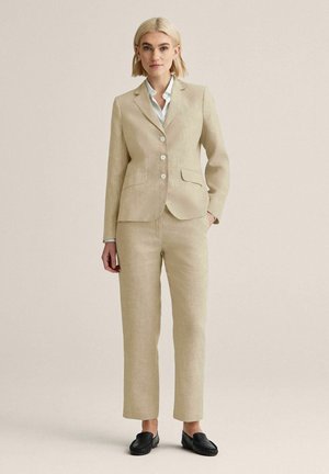 Kvinde stående iført en beige skræddersyet blazer og matchende bukser med en hvid skjorte og sorte loafers mod en neutral baggrund.