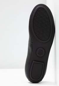 Semelle d’une chaussure en cuir noir avec grip texturé et logo circulaire, placée verticalement contre un mur et un sol blancs.