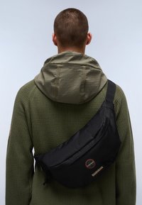 Borse a tracolla nera realizzata in tessuto resistente, con una superficie liscia, cinturino regolabile e una toppa del logo circolare sul fronte.
