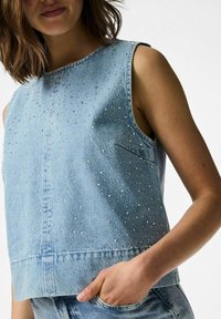 Svijetloplava traper bezrukavna majica s raspršenim rhinestones, okruglim izrezom i ravnim rubom. Uključuje detalj bočne džepove.