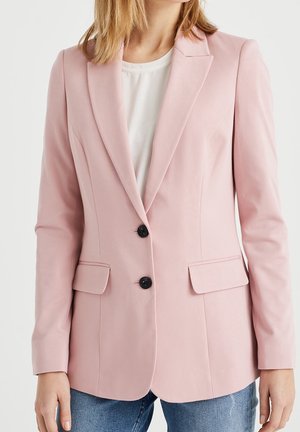 Femme portant un blazer ajusté rose clair avec des boutons noirs, des poches à rabat, une chemise blanche et un jean bleu sur un fond uni.