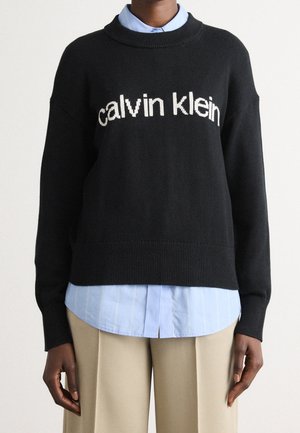 Personne portant un pull noir Calvin Klein par-dessus une chemise bleu clair à boutons et un pantalon beige sur fond blanc uni.