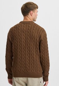 Brown knitted sweater featuring a cable pattern, ribbed cuffs and hem, with a classic crew neckline. The texture appears thick and cozy.
