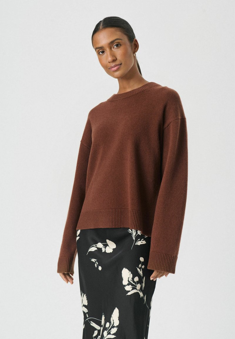 Suéter marrón de punto con cuello y dobladillo acanalados, con mangas largas. Llevado sobre una falda negra con estampado floral. Textura suave.