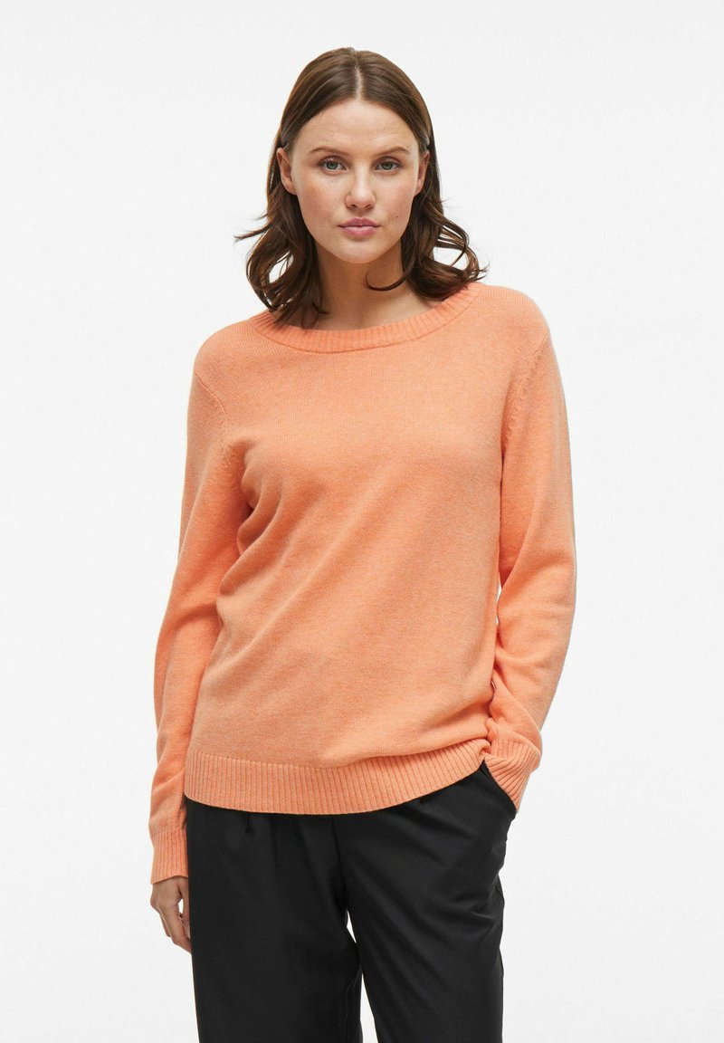 Maglione arancione chiaro con scollo rotondo, maniche lunghe e polsini e orlo a coste. Indossato con pantaloni neri, caratterizzati da una texture morbida.