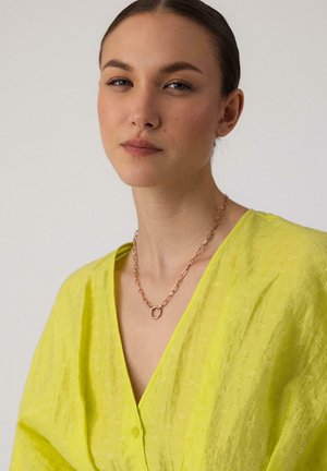 Junge Frau mit zurückgebundenen Haaren trägt eine leuchtend gelbe, strukturierte V-Ausschnitt-Bluse und eine Goldkette mit rundem Anhänger.
