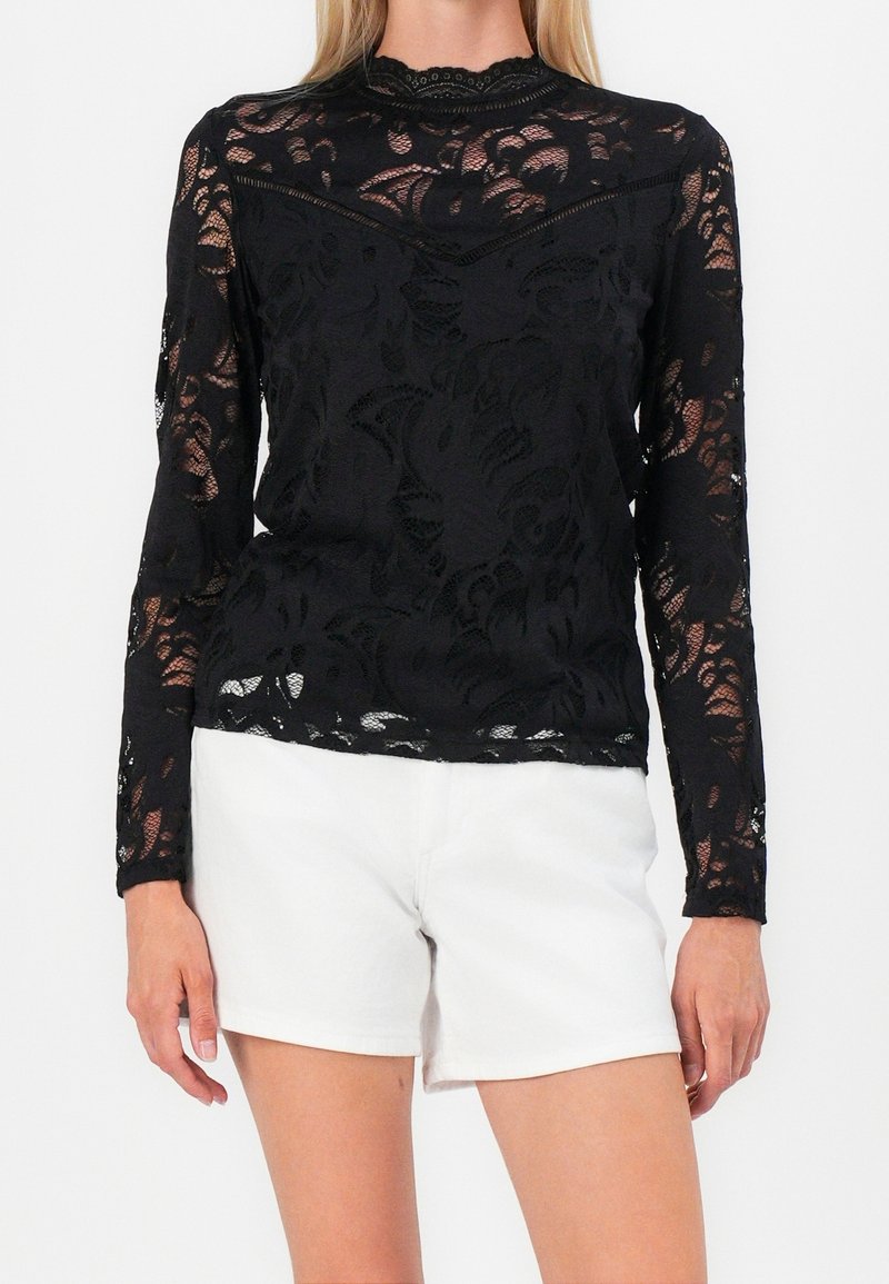 Femme portant un haut en dentelle noir à manches longues avec des motifs floraux et un short blanc, debout devant un fond uni.