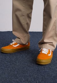 Orange suede sneakers with white accents, featuring a low-top design and rubber sole, paired with neutral-colored pants.