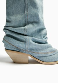 Denim-Ankle-Boot mit einem lässigen Design, hellblauer Farbe, beigem gestreiften Absatz, sichtbaren Nähten und abgerundeter Zehenkappe.