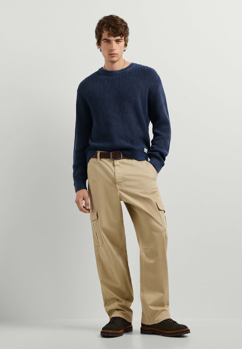 Jeune homme debout portant un pull en maille bleu marine, un pantalon cargo beige avec poches latérales, une ceinture marron et des chaussures marron foncé sur un fond uni.