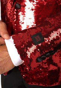 Roka, kas tur baltā krekla piedurkni zem sarkana spīdīgu mērci ar melnām pogām un Oppo Suits etiķeti.