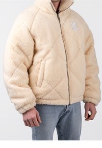 Crème-kleurige fleece jas met een ruitpatroon, ritssluiting aan de voorkant en zijzakken. Beschikt over een logo op de linkerkant van de borst en ribgebreide manchetten.