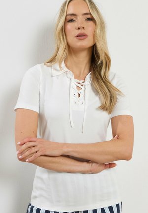 Femme blonde aux cheveux lâchés portant un polo blanc à lacets et un pantalon rayé, bras croisés, debout contre un mur clair uni.