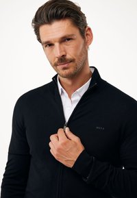 Černý pletený svetr s vysokým límcem a zapínáním na zip, který má decentní logo na hrudi a žebrované manžety pro přidanou strukturu.