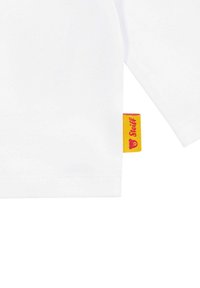 Camicia bianca di cotone a maniche lunghe, con un piccolo etichetta gialla con scritto rosso sul lato. Texture liscia, design semplice.