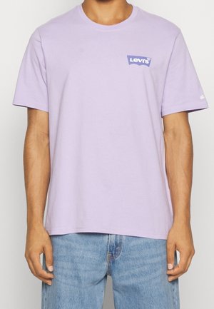 Mann trägt ein hellviolettes T-Shirt von Levi's mit Logo auf der linken Brust, kombiniert mit hellblauen Jeans, steht vor einem einfarbigen Hintergrund.