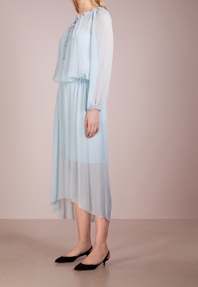 Femme debout portant une robe midi bleu clair à manches longues avec taille élastique et chaussures slingback noires à talons bas pointues.